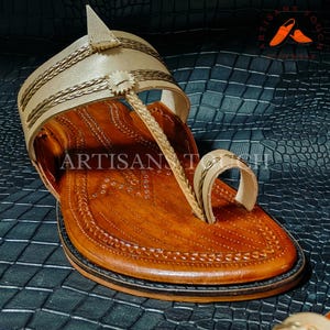 May include: Handmade leather sandal in a warm brown hue with intricate stitching and a cream-colored strap. The sandal features a decorative braided design and the text "ARTISAN TOUCH". The sole is dark brown, and the background is a textured dark blue.