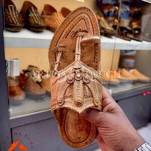 May include: A light brown leather sandal with intricate carved details. The sandal features a rounded toe and a strap that goes between the toes. The words "ARTISANS TOUCH FOOTWEAR" are visible in the image.