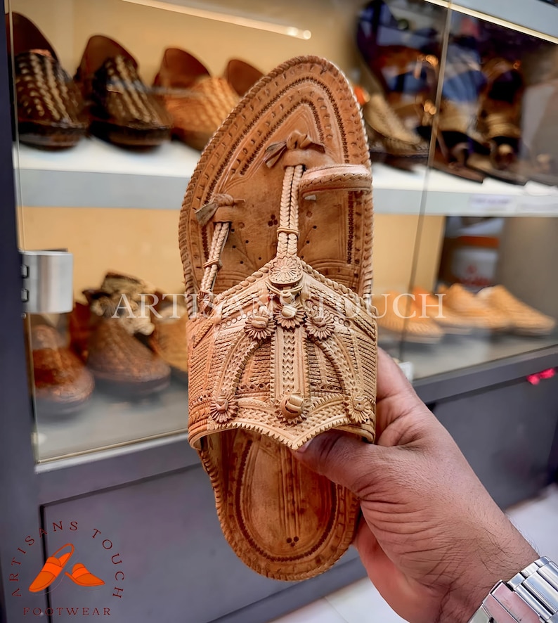 May include: A light brown leather sandal with intricate carved details. The sandal features a rounded toe and a strap that goes between the toes. The words "ARTISANS TOUCH FOOTWEAR" are visible in the image.