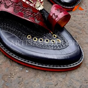 May include: Close-up of a black leather sandal with intricate embossed floral patterns and a red accent. The sandal features gold eyelets and a red sole. The text "ARTISAN TOUCH FOOTWEAR" is visible.