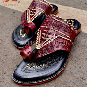 May include: Pair of handcrafted leather sandals with intricate floral and geometric patterns. The sandals feature a deep burgundy upper with gold accents and a black sole. The toe strap and upper are adorned with decorative stitching and metal studs. The sole has a red trim.