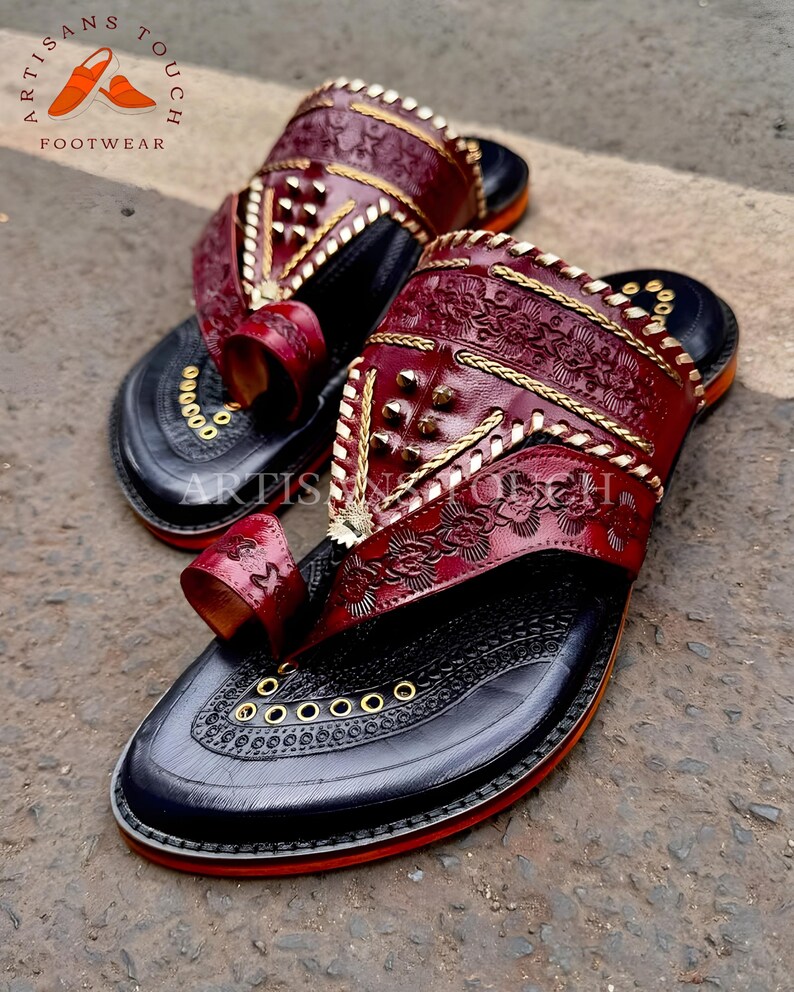 May include: Pair of handcrafted leather sandals with intricate floral and geometric patterns. The sandals feature a deep burgundy upper with gold accents and a black sole. The toe strap and upper are adorned with decorative stitching and metal studs. The sole has a red trim.