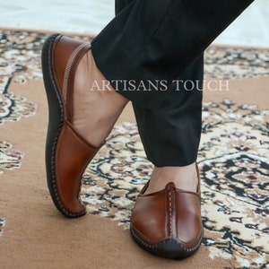 May include: Brown leather slip-on shoes with a rounded toe and stitched detailing. The shoes have a dark sole and are worn with black trousers. The text "ARTISANS TOUCH" is visible on the image.