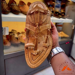 May include: Hand-carved, light brown leather sandal with intricate floral and geometric designs. The sandal features a detailed strap and sole, showcasing artisanal craftsmanship. The image also displays other footwear in the background.