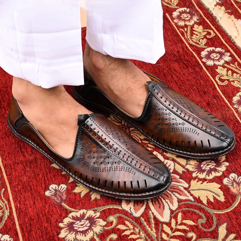 K&ouml;nnte beinhalten: Ein Paar braune Lederschlappen mit perforiertem Design. Die Schuhe haben eine spitze Zehenpartie und einen niedrigen Absatz.