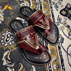 May include: A pair of handcrafted leather sandals with a dark red upper and black sole. The sandals feature intricate gold stitching and decorative metal studs. The design includes a toe loop and a strap across the top of the foot.