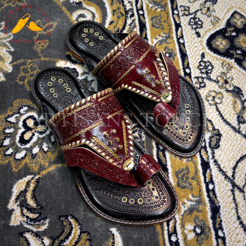 May include: A pair of handcrafted leather sandals with a dark red upper and black sole. The sandals feature intricate gold stitching and decorative metal studs. The design includes a toe loop and a strap across the top of the foot.