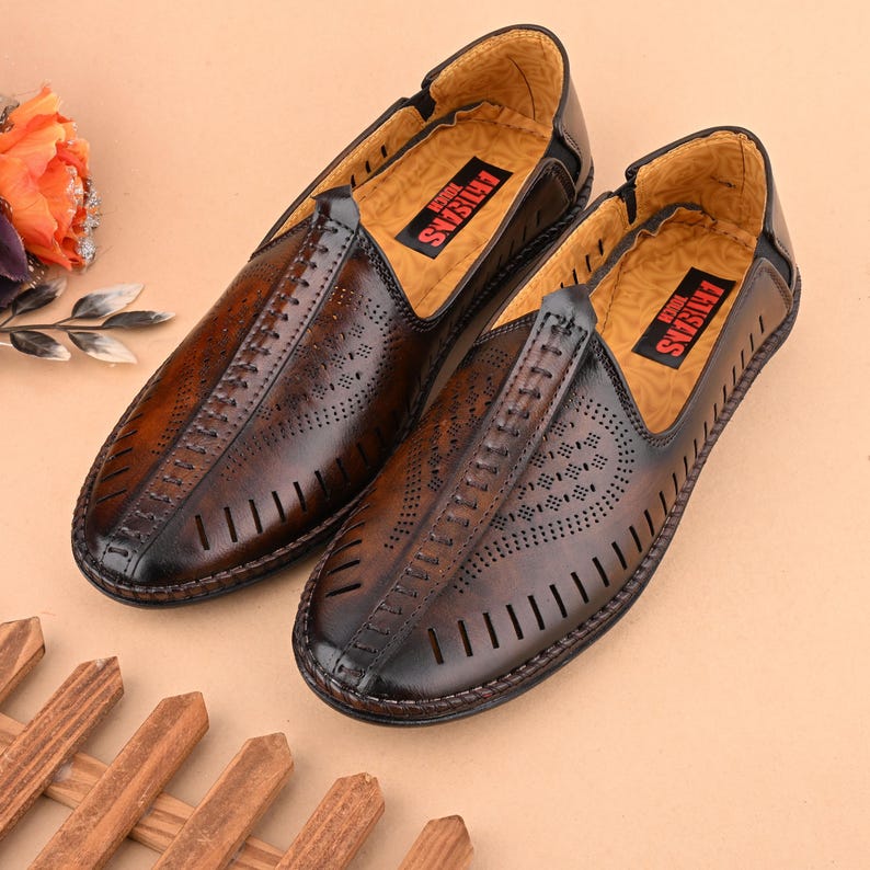 K&ouml;nnte beinhalten: Ein Paar braune Lederschuhe zum Hineinschl&uuml;pfen mit ausgeschnittenen Designs und dem Text "FOR ARTISANS" auf der Innensohle.