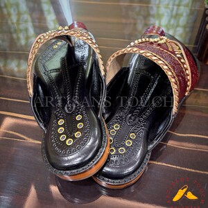 May include: A pair of black leather sandals with intricate stitching and gold accents. The sandals feature a wooden sole and a decorative strap with gold trim. The text "ARTISANS TOUCH" is visible on the sandals.