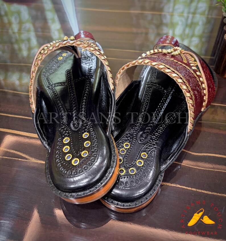 May include: A pair of black leather sandals with intricate stitching and gold accents. The sandals feature a wooden sole and a decorative strap with gold trim. The text "ARTISANS TOUCH" is visible on the sandals.