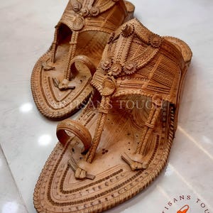 May include: A pair of intricately carved, light brown leather sandals. The sandals feature detailed patterns, including floral and geometric designs, with a toe loop and ankle strap. The footwear is displayed on a light-colored surface.