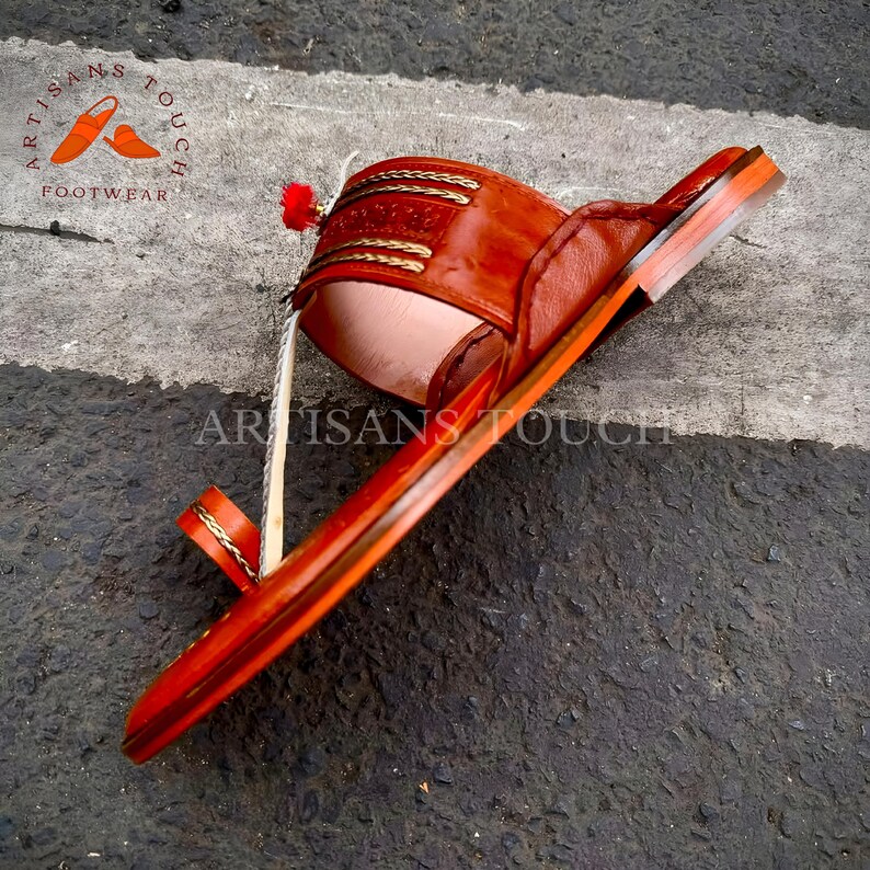 May include: A handcrafted leather sandal in shades of brown and orange. The sandal features intricate stitching and a decorative tassel. The sole is made of wood. The text "ARTISANS TOUCH FOOTWEAR" is visible.