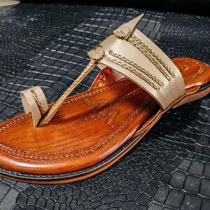May include: A handcrafted leather sandal in shades of tan and brown. The sandal features a toe loop and a strap adorned with gold-colored accents. The sole is a darker brown, and the sandal is set against a textured black surface.