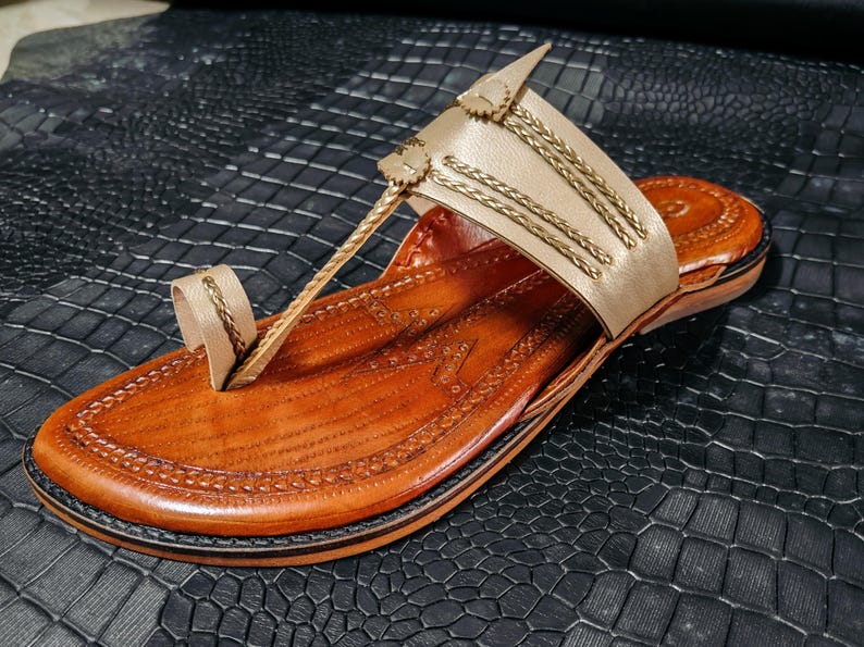 May include: A handcrafted leather sandal in shades of tan and brown. The sandal features a toe loop and a strap adorned with gold-colored accents. The sole is a darker brown, and the sandal is set against a textured black surface.
