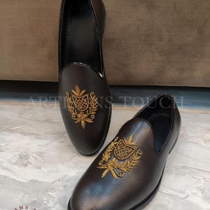 Op de afbeelding: Een paar bruinleren instappers met gouden geborduurde wapenversieringen. De schoenen hebben een ronde neus en een zwarte zool. De merknaam "ARTISANS TOUCH FOOTWEAR" is linksonder zichtbaar.