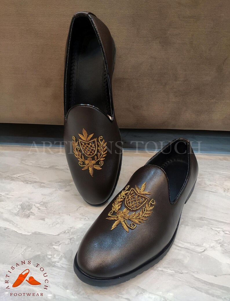 Op de afbeelding: Een paar bruinleren instappers met gouden geborduurde wapenversieringen. De schoenen hebben een ronde neus en een zwarte zool. De merknaam "ARTISANS TOUCH FOOTWEAR" is linksonder zichtbaar.