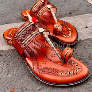 May include: A pair of handcrafted, orange leather sandals with intricate embossed patterns. The sandals feature a toe loop, decorative stitching, and small red pom-poms. The sole is adorned with brass eyelets. The sandals are labeled "Artisans Touch Footwear".