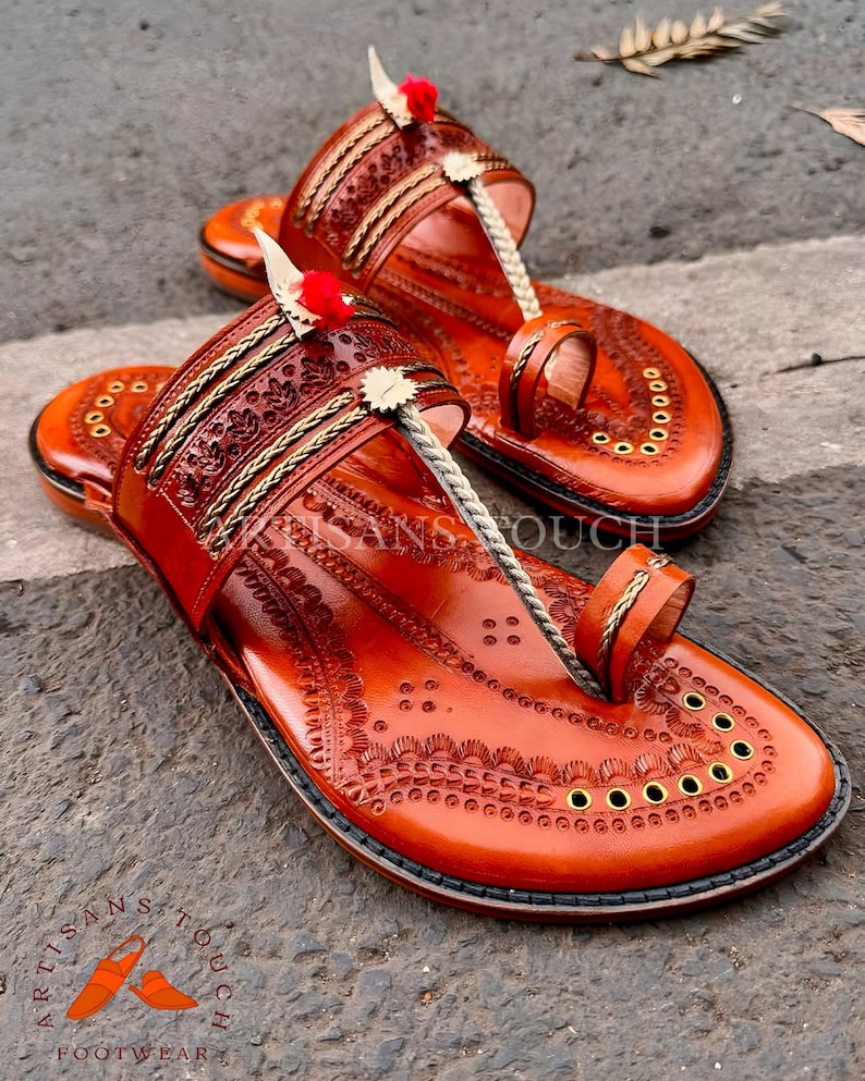 May include: A pair of handcrafted, orange leather sandals with intricate embossed patterns. The sandals feature a toe loop, decorative stitching, and small red pom-poms. The sole is adorned with brass eyelets. The sandals are labeled "Artisans Touch Footwear".