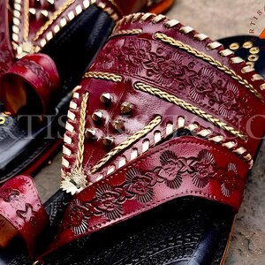 May include: Close-up of handcrafted leather sandals. The sandals feature a deep red, embossed leather design with gold accents and studded details. The soles are black with gold embellishments. The text "ARTISANS TOUCH FOOTWEAR" is visible.
