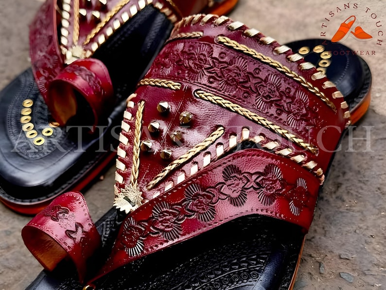 May include: Close-up of handcrafted leather sandals. The sandals feature a deep red, embossed leather design with gold accents and studded details. The soles are black with gold embellishments. The text "ARTISANS TOUCH FOOTWEAR" is visible.