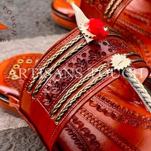 May include: Close-up of handcrafted leather sandals in a warm reddish-brown hue. The sandals feature intricate embossed patterns, gold-colored braided trim, and decorative elements. The sandals have a toe strap and a heel strap. The brand name "ARTISANS TOUCH FOOTWEAR" is visible.