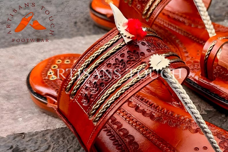 May include: Close-up of handcrafted leather sandals in a warm reddish-brown hue. The sandals feature intricate embossed patterns, gold-colored braided trim, and decorative elements. The sandals have a toe strap and a heel strap. The brand name "ARTISANS TOUCH FOOTWEAR" is visible.