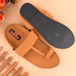Op de afbeelding: Een paar bruine leren sandalen met een band over de bovenkant van de voet en een teenlus. De sandalen hebben een zwarte zool en de tekst "TOUGH ARTISANS" is op de band gedrukt.
