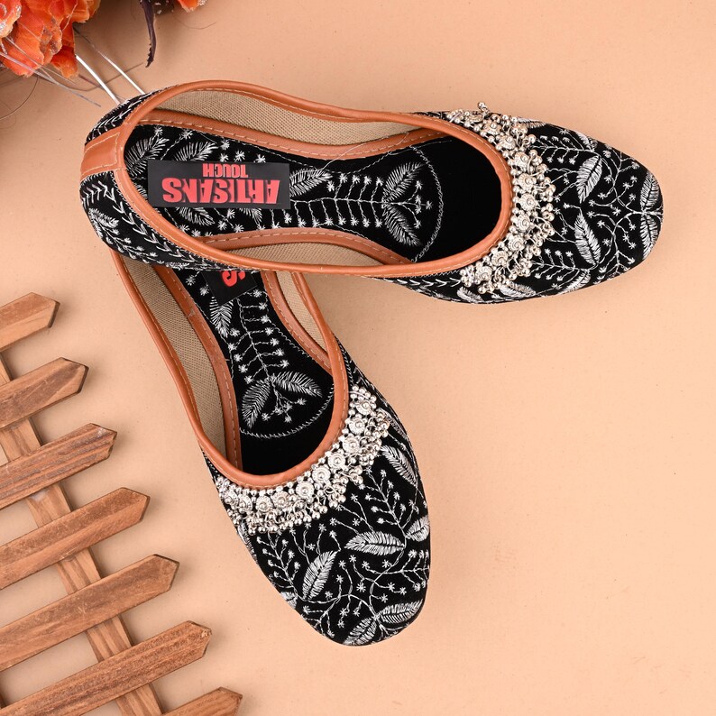 May include: Black velvet embroidered flat shoes with silver embellishments. The shoes have a brown leather insole with the text "ARTISANS TOUCH" printed on it.