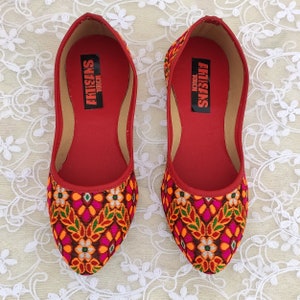 May include: A pair of red flat shoes with a floral pattern. The shoes have a pointed toe and are made of a fabric with a woven design. The inside of the shoes is red and has the text "ARTISANS TOUCH" printed on it.