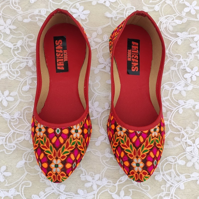 May include: A pair of red flat shoes with a floral pattern. The shoes have a pointed toe and are made of a fabric with a woven design. The inside of the shoes is red and has the text "ARTISANS TOUCH" printed on it.