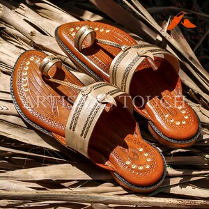 May include: A pair of handcrafted brown leather sandals with beige straps and intricate stitching. The sandals feature a toe loop and a decorative design on the footbed. The image includes the text "ARTISANS TOUCH FOOTWEAR".
