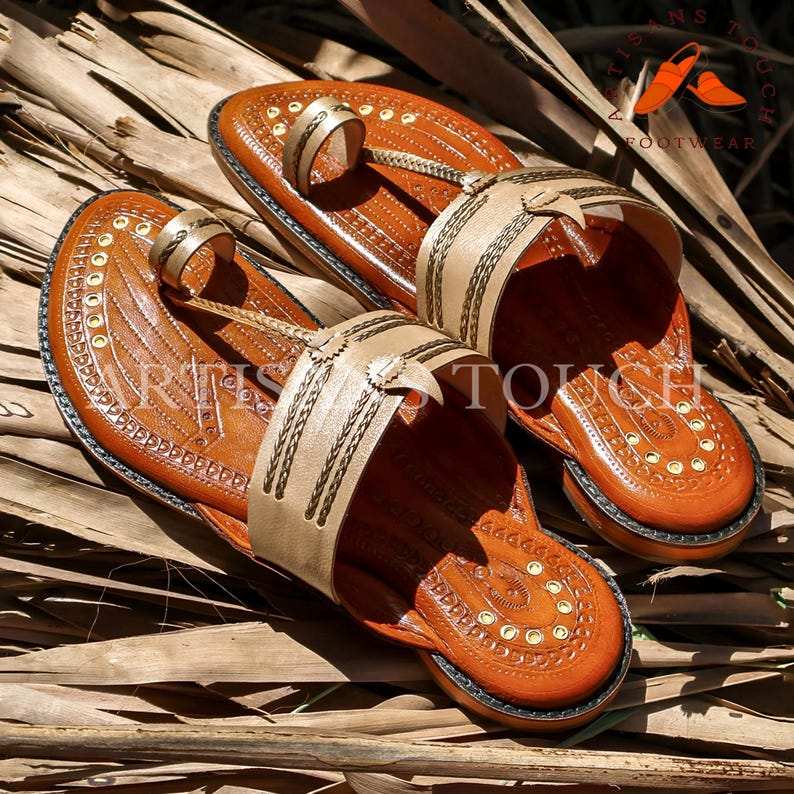 May include: A pair of handcrafted brown leather sandals with beige straps and intricate stitching. The sandals feature a toe loop and a decorative design on the footbed. The image includes the text "ARTISANS TOUCH FOOTWEAR".