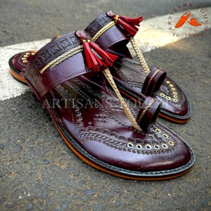 May include: Burgundy leather sandals with intricate embossed patterns and gold accents. The sandals feature a toe loop, decorative tassels, and the text "ARTISANS TOUCH FOOTWEAR". The soles have a contrasting orange trim.
