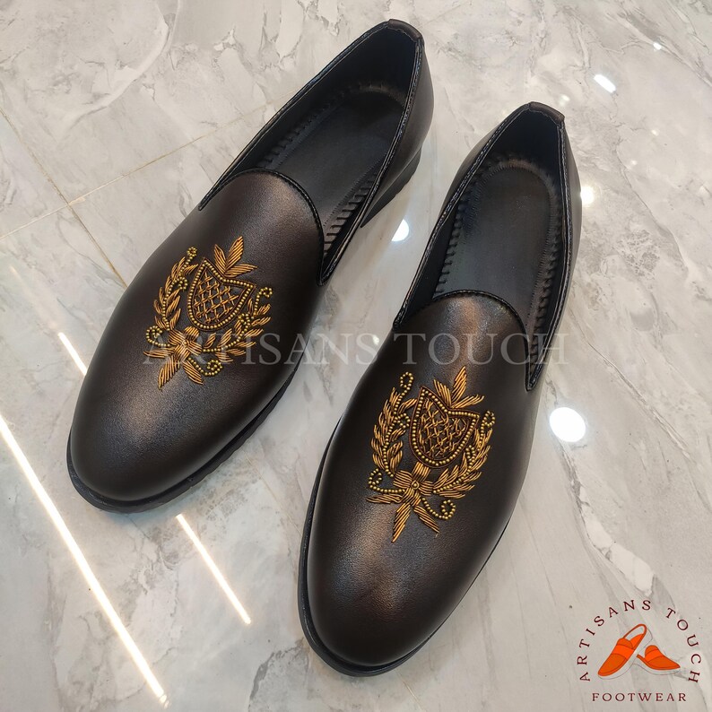 Op de afbeelding: Een paar donkerbruine leren loafers met goud geborduurde wapenontwerpen. De schoenen hebben een ronde neus en een lage hak. De tekst "ARTISANS TOUCH FOOTWEAR" is rechtsonder zichtbaar.