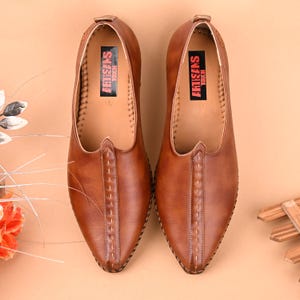 May include: A pair of brown leather slip-on shoes with pointed toes. The shoes have a stitched design down the center and a red label inside that reads "ARTISANS TOUCH". The shoes are displayed on a light brown surface.