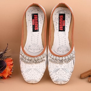 May include: White embroidered flat shoes with silver embellishments and a brown leather insole. The insole has the text "ARTISANS TOUCH" printed on it.