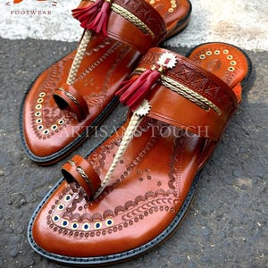 May include: A pair of brown leather sandals with intricate embossed patterns and decorative elements. The sandals feature a toe loop, a strap across the top, and a red tassel. The sandals are labeled "ARTISANS TOUCH FOOTWEAR".