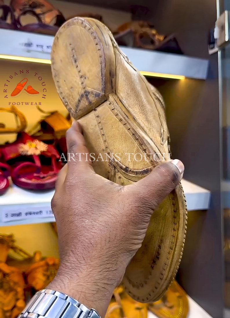 May include: Close-up of a tan leather sandal sole. The sandal is held up, showcasing the textured surface and stitching details. The logo "ARTISANS TOUCH FOOTWEAR" is visible. Other sandals are on display in the background.