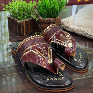 May include: Burgundy leather sandals with gold accents and intricate embossed patterns. The sandals have a black sole and a toe loop design. The image also shows the text "ARTISANS TOUCH FOOTWEAR".