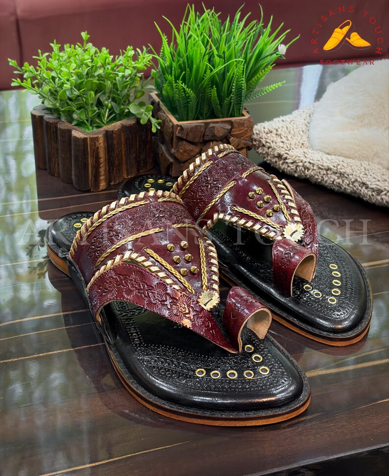 May include: Burgundy leather sandals with gold accents and intricate embossed patterns. The sandals have a black sole and a toe loop design. The image also shows the text "ARTISANS TOUCH FOOTWEAR".