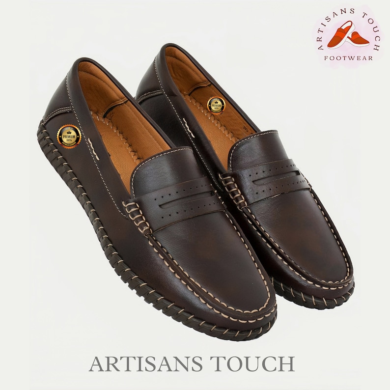May include: A pair of brown leather loafers with a stitched edge and a decorative stitch detail on the vamp. The loafers have a leather sole and a comfortable fit. The brand name "Artisans Touch Footwear" is printed on the image.