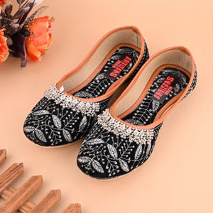 May include: A pair of black embroidered flat shoes with silver embellishments. The shoes have a brown leather insole and the text "ARTISANS TOUCH" is printed on the inside of each shoe.