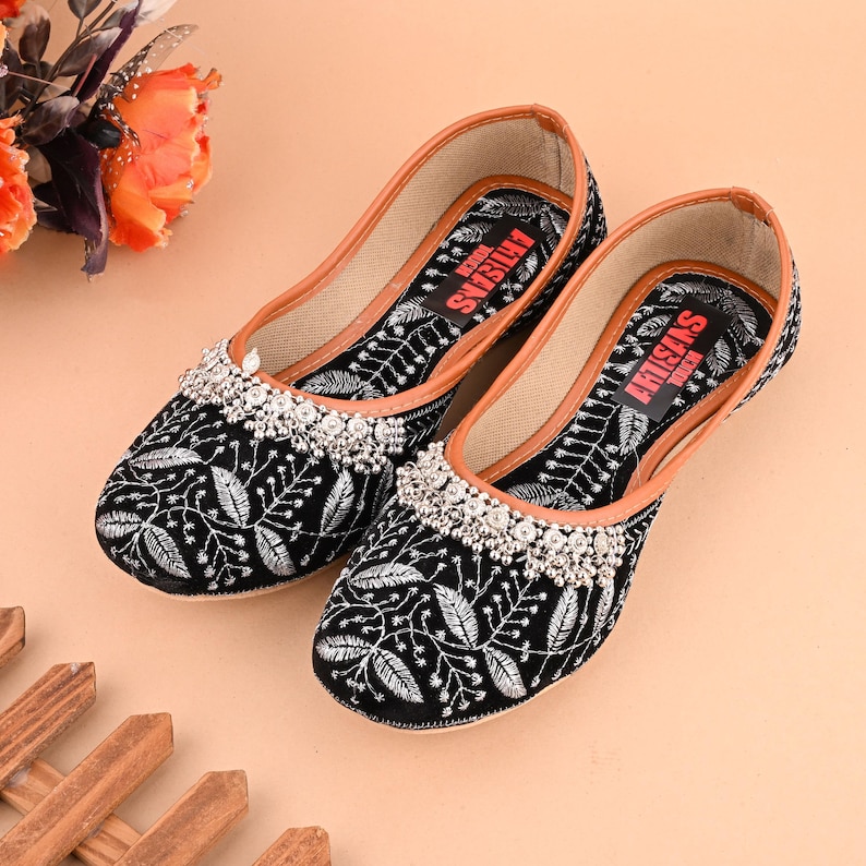 May include: A pair of black embroidered flat shoes with silver embellishments. The shoes have a brown leather insole and the text "ARTISANS TOUCH" is printed on the inside of each shoe.