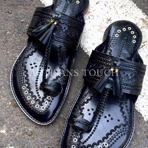 May include: A pair of black leather sandals with intricate embossed patterns and decorative stitching. The sandals feature a toe loop, a strap across the top, and small tassels. The edges are trimmed with white and adorned with gold and blue accents. The sandals are labeled "ARTISANS TOUCH FOOTWEAR".