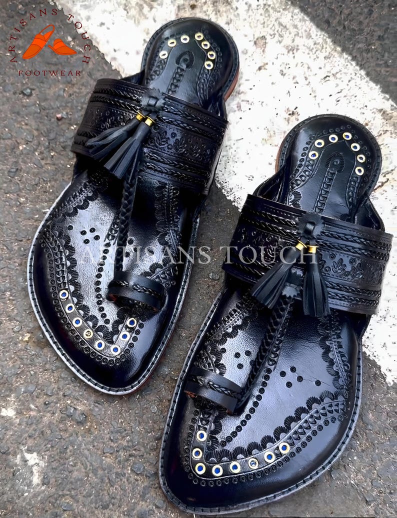 May include: A pair of black leather sandals with intricate embossed patterns and decorative stitching. The sandals feature a toe loop, a strap across the top, and small tassels. The edges are trimmed with white and adorned with gold and blue accents. The sandals are labeled "ARTISANS TOUCH FOOTWEAR".