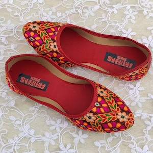 May include: A pair of red embroidered flat shoes with a floral pattern. The shoes have a red insole with the text "ARTISANS TOUCH" printed on them.