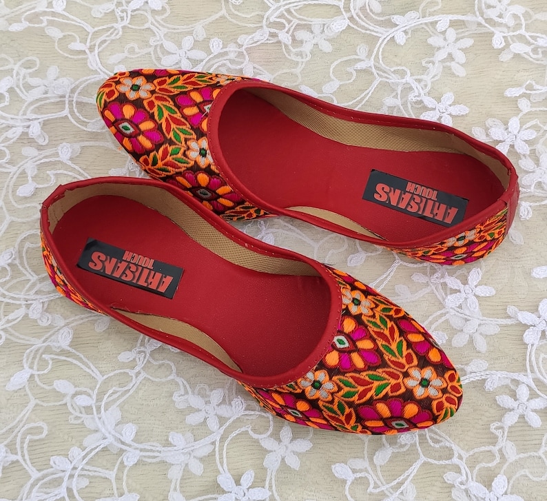 May include: A pair of red embroidered flat shoes with a floral pattern. The shoes have a red insole with the text "ARTISANS TOUCH" printed on them.