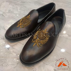 Op de afbeelding: Een paar bruine leren loafers met goud geborduurde wapenontwerpen. De schoenen hebben een ronde neus en een lage hak. De tekst "ARTISAN TOUCH" is zichtbaar.