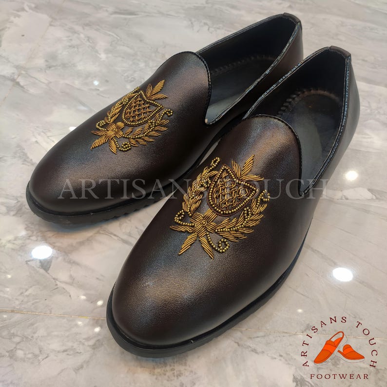 Op de afbeelding: Een paar bruine leren loafers met goud geborduurde wapenontwerpen. De schoenen hebben een ronde neus en een lage hak. De tekst "ARTISAN TOUCH" is zichtbaar.