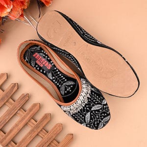 May include: Black velvet embroidered flat shoes with silver embellishments. The shoes have a brown leather insole with the text "ARTISANS" and "FASHION" printed on them.